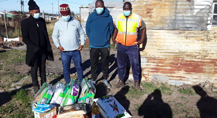 Four masked men stand before a large pile of food, showcasing a moment of community and sharing amidst the abundance.